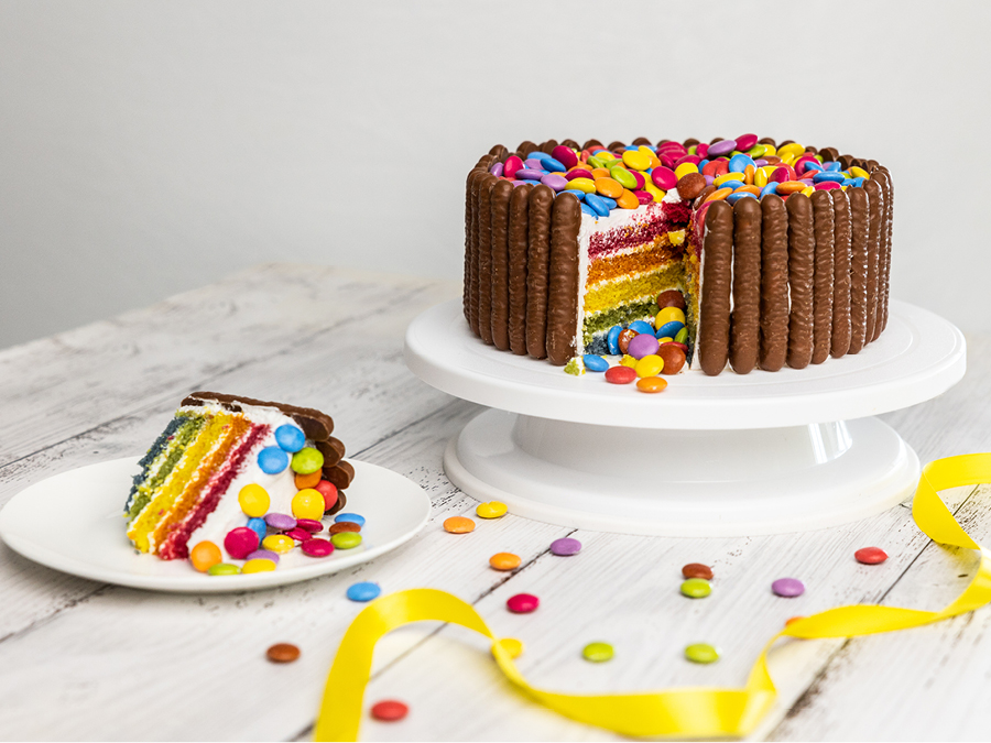 A store bought rainbow cake covered in smarties with chocolate fingers lined up around the side. The cake sits on a white cake stand and has a slice cut out.