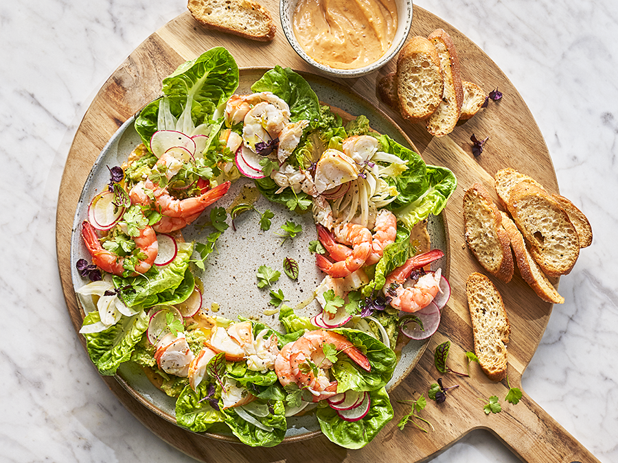 A prawn cocktail recipe displayed in the shape of a Christmas wreath using lettuce, prawns, avocado and chipotle mayo. Served on a round wooden chopping board with slices of bread.