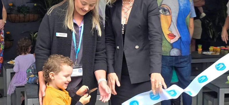 A child cuts a ribbon with scissors during a ceremony, flanked by two adults. The ribbon features blue logos. In the background, several people are present, including one wearing a colorful shirt with an abstract face design. The event appears to be indoors, decorated with plants.