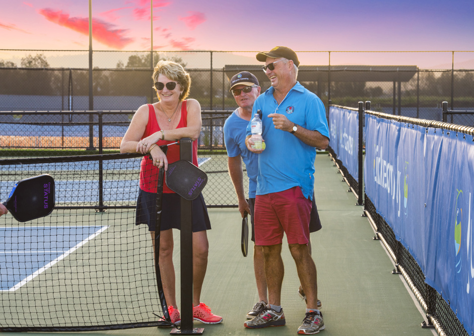 Group of three home owners standing on a Halcyon Greens pickleball court near the net while laughing and smiling 
