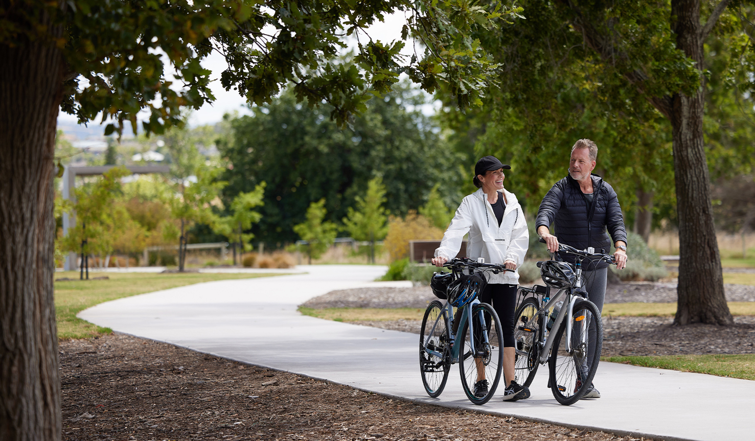 A man and woman walking next to their bikes along a path surrounded by trees.