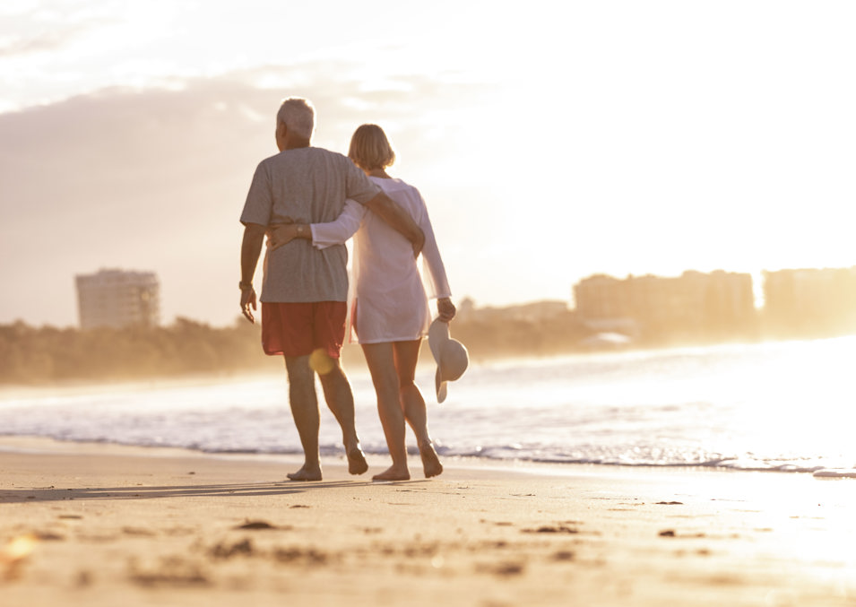 A man and woman walk leisurely with their arms around each other at sunrise on the Mooloolaba beach shoreline.,