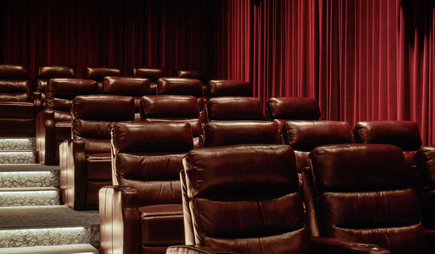 Indoor gold-class style luxurious cinema with dark red leather recliner chairs tiered up stairs with velvet red curtains lining the walls and intimate lighting.