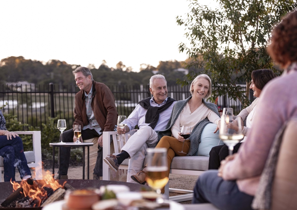 A group of adults in their sixties gather around an outdoor fireplace with glasses of white wine in hand in the afternoon.