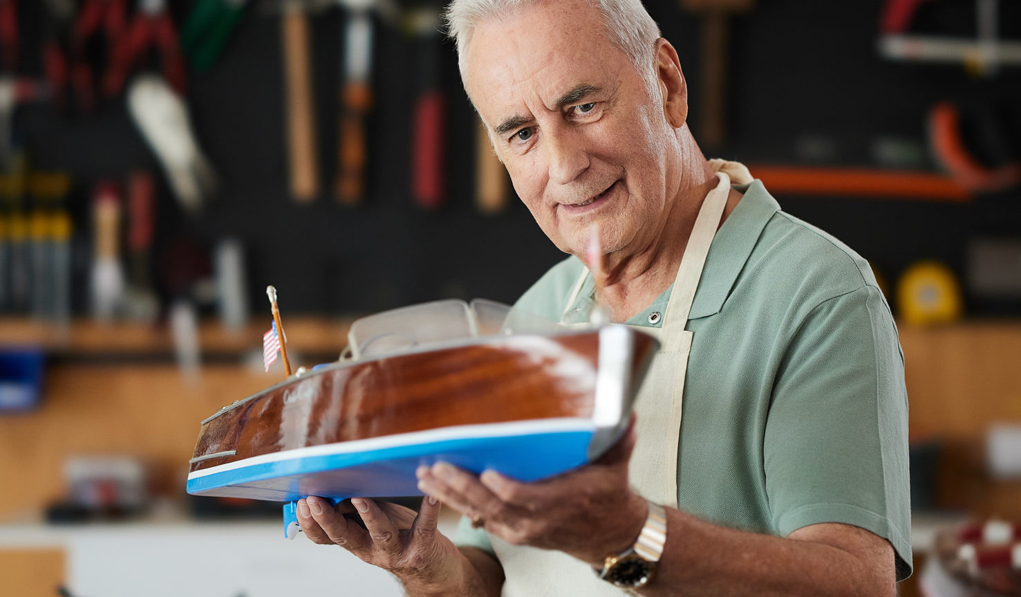 A man in his sixties hold a timber model boat whilst wearing a cream apron and green polo shirt in a wood work shop.