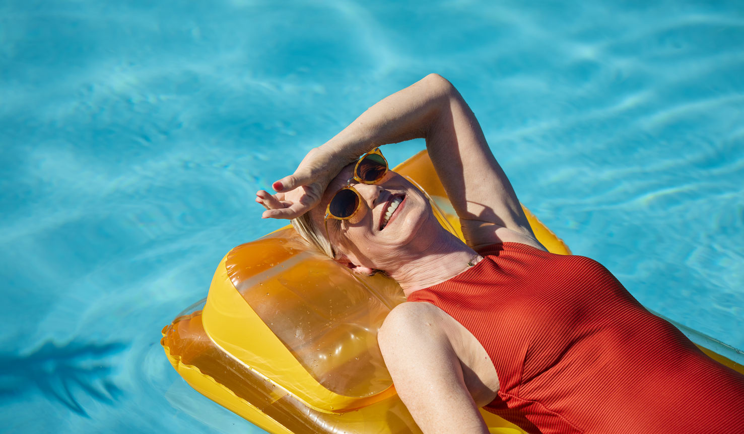 A woman in his sixties lounges in a blue pool on a yellow inflatable sun lounger whilst wearing a red one piece swimsuit and yellow sunglasses.