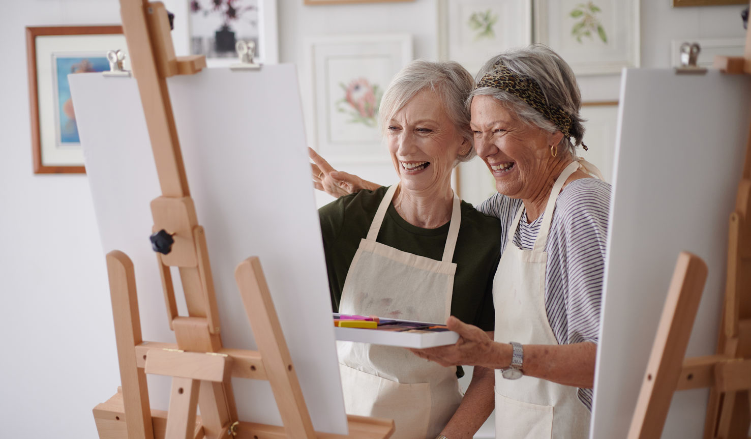 Two women in their sixties stand in front of two painters easels whilst holding crayons in a craft room,