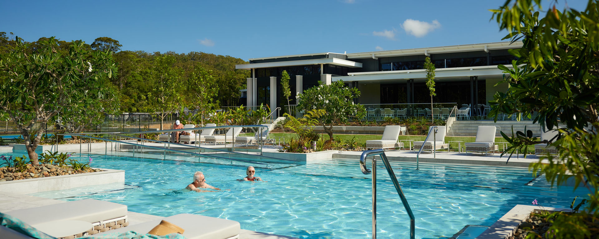 An outdoor resort pool with lush tropical landscaping in front of a modern recreational precinct building.