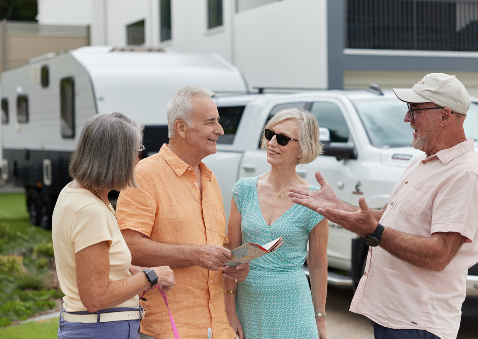 Four adults in their sixties laugh and talk together in a group in front of a house with side access and a large car towing a caravan in a lifestyle living community.