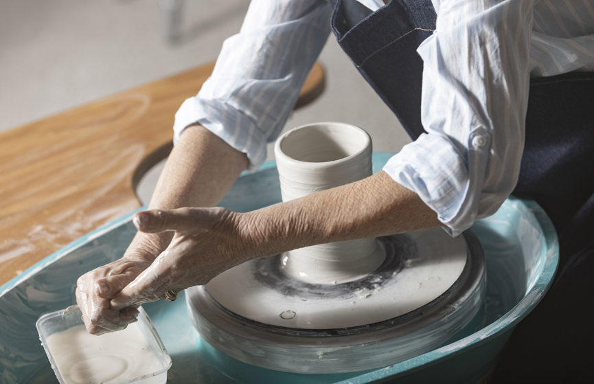 A woman's hands mould a clay pot over a pottery wheel whilst wearing a blue apron and light blue striped button up shirt.