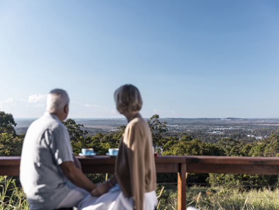 A man and woman sit side by side on stools perched on a timber balcony with a cup of coffee overlooking the Sunshine Coast hinterland on a clear day.