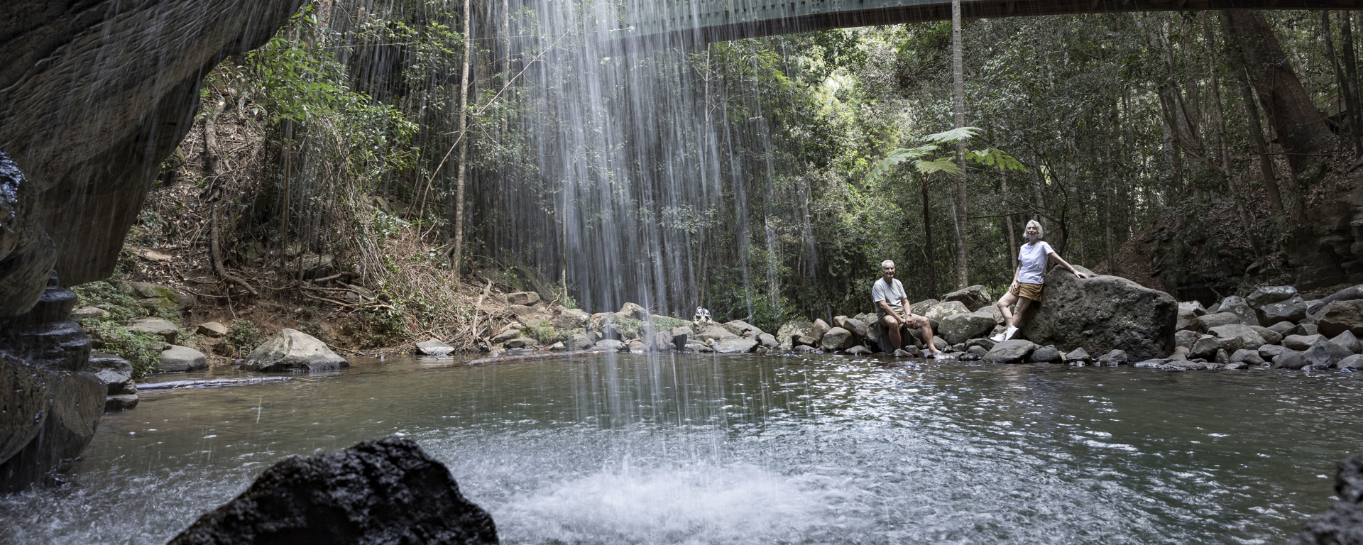 A man and woman relax by the base of a waterfall within the Buderim rainforest underneath a bridge on a cloudy day.