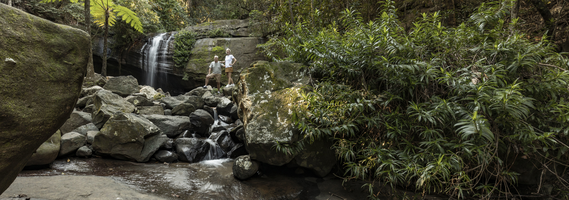 A man and woman relax a top a rocky area in front of a waterfall surrounded by a tropical rainforest.