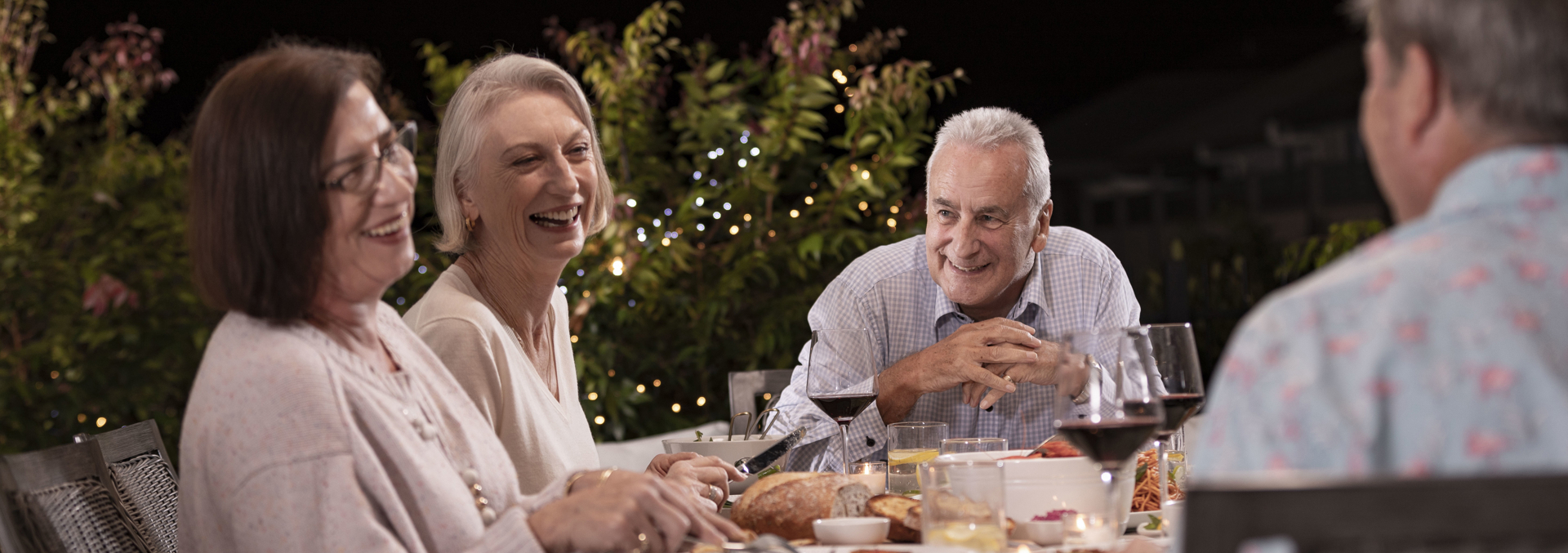 A group of four adult friends smile and laugh at each other whilst enjoying a glass of wine and dinner meal at night time.