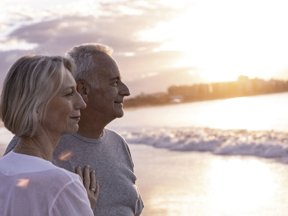 A man and woman stand close to each other in an embrace overlooking the ocean at sunset.