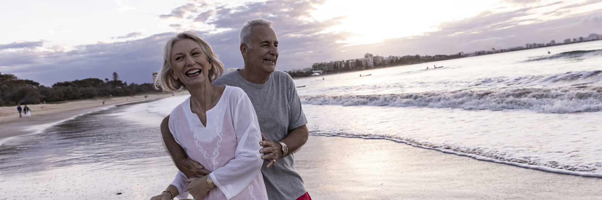 A man and woman embrace playfully overlooking the ocean at sunset whilst wearing summer clothing.