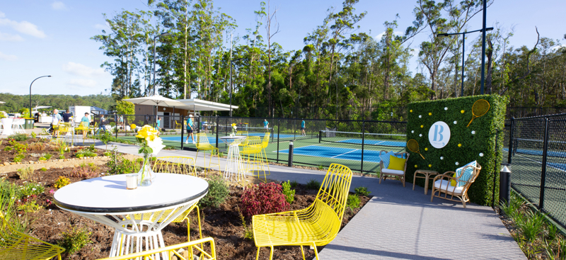 An outdoor sports pavilion area with four pickleball courts and tennis court, adorned with yellow and white tables and chairs with a green media wall sirtuated in the middle.