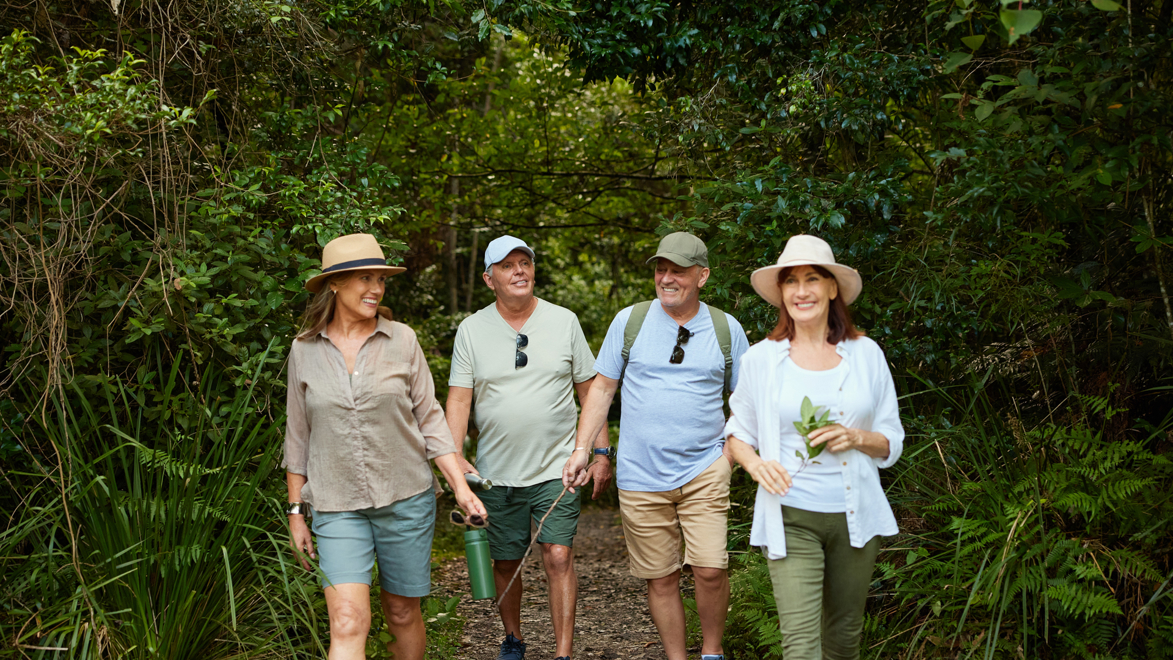 a group of seniors over 50 walk together in a green forest wearing casual walking gear and smiling at each other