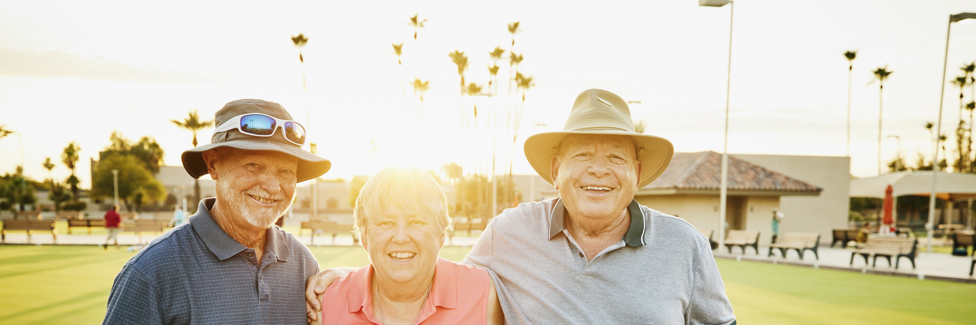 a group of three over 50s stand together embracing around the shoulder in front of a bowling green at sunset