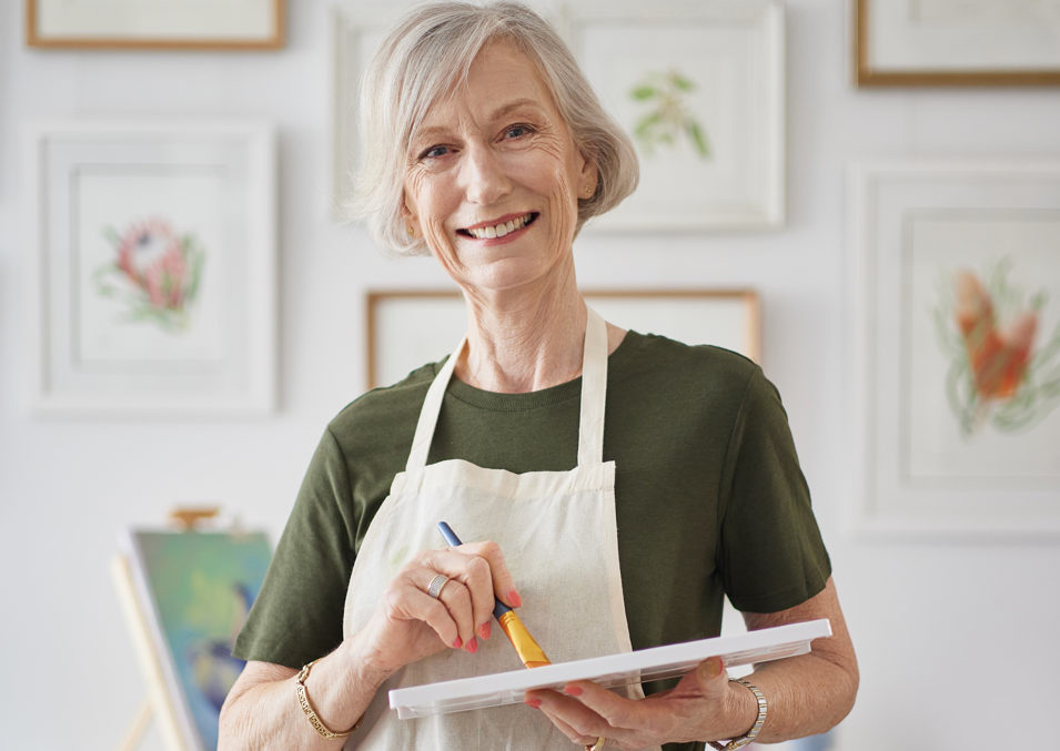 A woman in her sixties wearing a white apron and holding a painting pallete