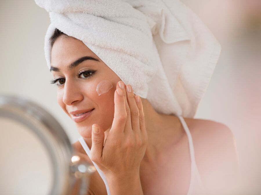 A Woman with her hair wrapped up in a towel applying lotion to her cheek