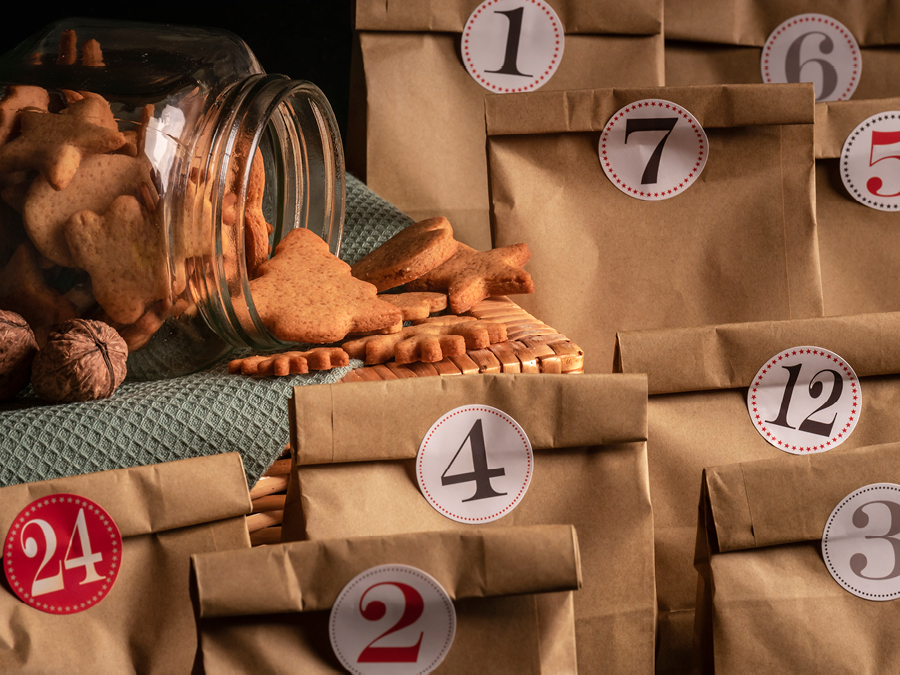 9 brown paper bags arranged around a jar of cookies with numbered stickers on them indicating which day of December they should be opened