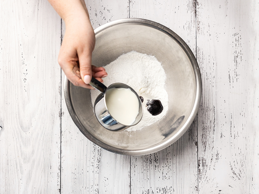 A metal mixing bowl with powdered icing and vanilla paste, with a cup of milk being poured in. 