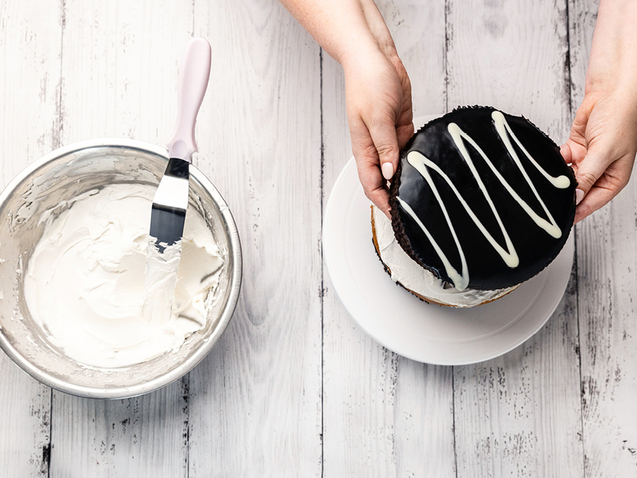 A metal mixing bowl filled with cake icing sitting on a white wooden benchtop. A chocolate mud cake is sitting on a plate ready to be iced. 
