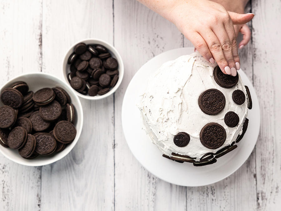 A person's hands placing Oreo cookies on top of an iced mud cake. Two bowls of different sized Oreo cookies are placed beside the cake. 