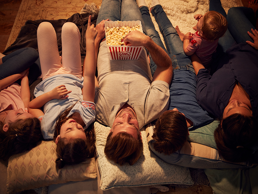 A family of six sitting on the couch with popcorn watching a movie.