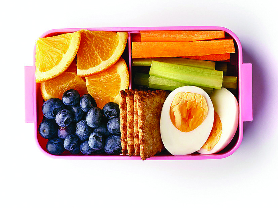 A pink lunchbox with chopped vegetables and fruit including carrots, celery, orange and grapes, with some crackers and boiled eggs.