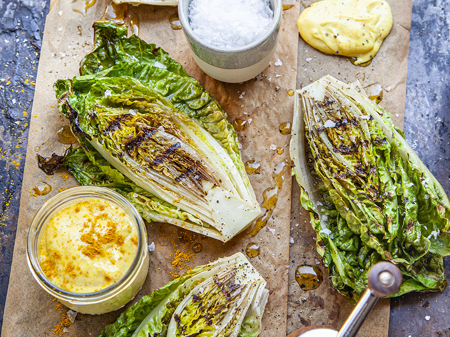 Grilled romaine lettuce served on a wooden chopping board with a side of curry mayonnaise in a glass jar.