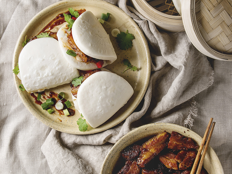 Three pork belly bao buns served on a taupe stone plate next to a bowl of cooked pork belly slices. 