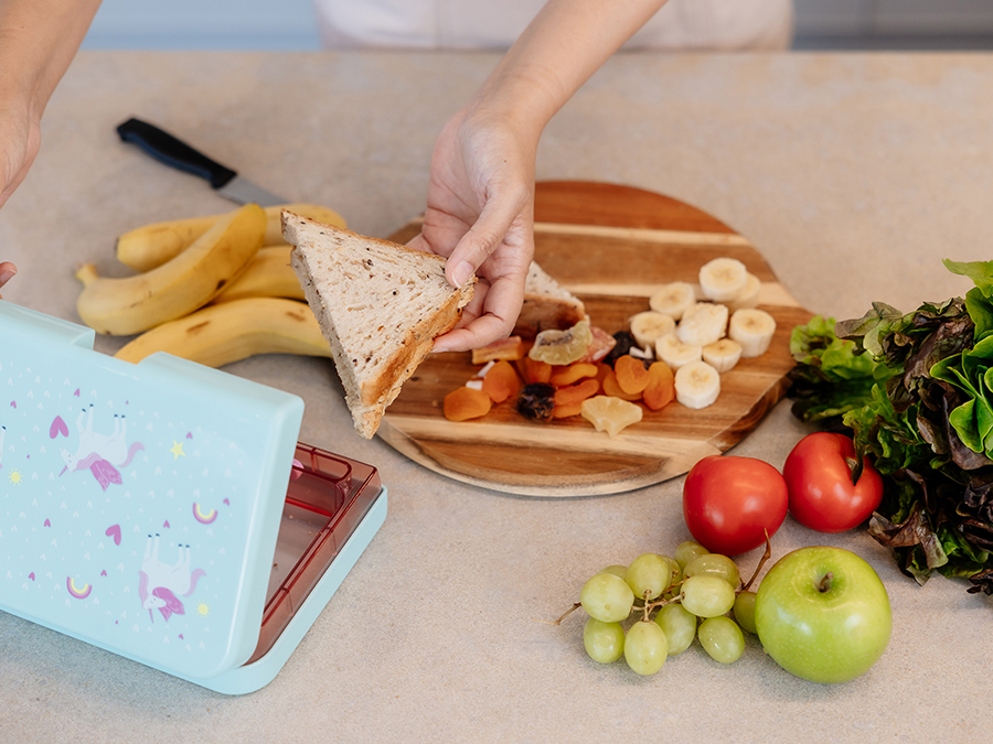 A kitchen bench with a wooden chopping board with chopped fruit and a triangular sliced sandwich. The Sandwich is being placed inside a blue kids lunchbox. 