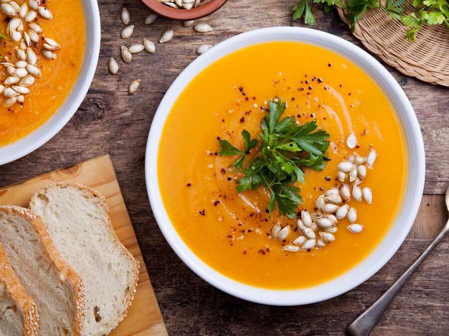 Pumpkin soup topped with coriander and pumpkin seeds served in a white bowl with a side of sliced bread. 
