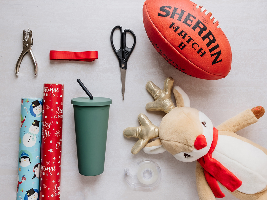 A flatlay of various items for gift wrapping including Christmas gift wrapping paper, scissors, ribbon, a rugby and reusable smoothie cup
