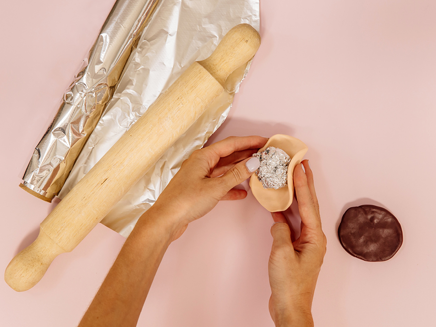 Hands sculpting clay into preferred shape with rolling pin and aluminium foil displayed on table.